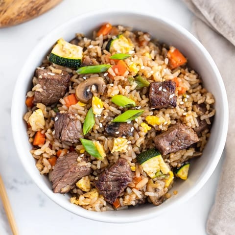 Golden-brown cubes of tender steak nestled in a savory mix of fried rice, vibrant peas, and diced vegetables in a hot skillet.  